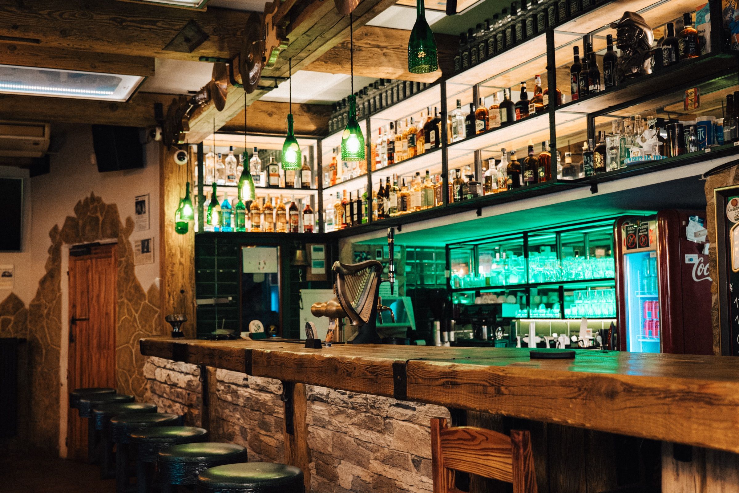 Dlouhý dřevěný bar se zeleně podsvícenými policemi a výčepem Guinness