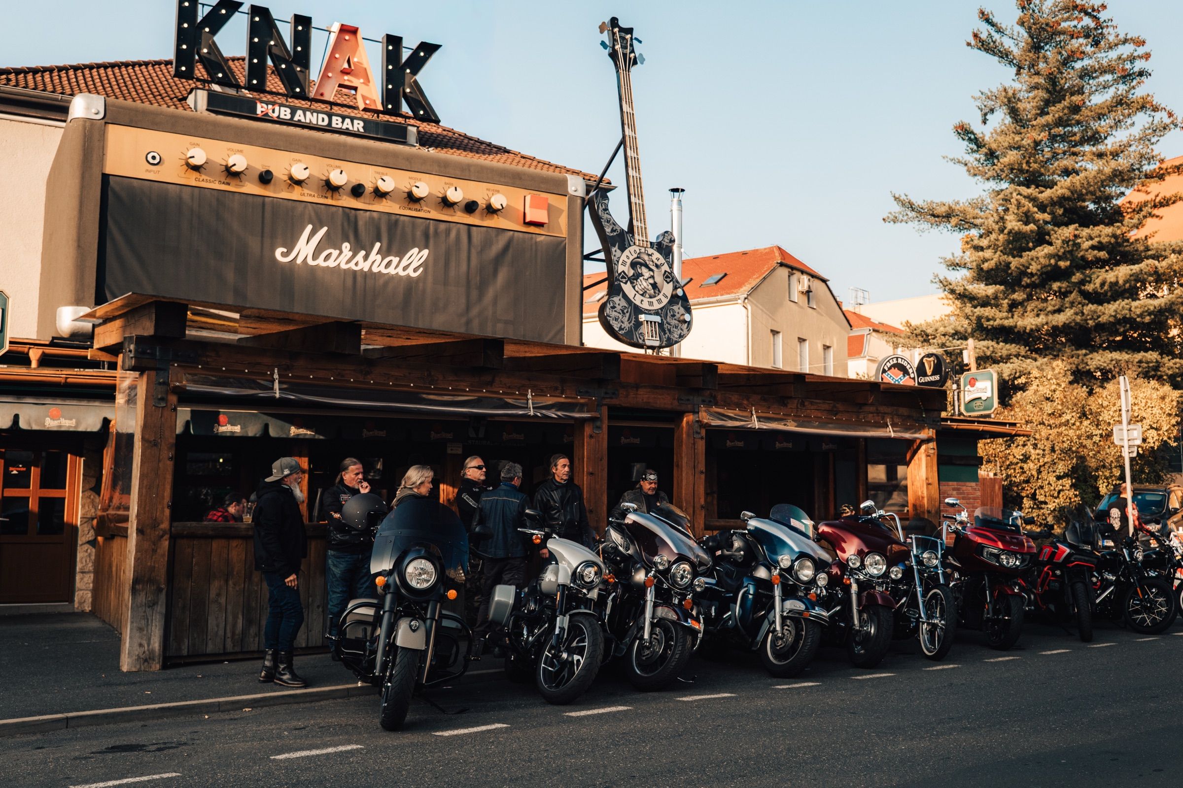 Řada motorek zaparkovaná podél dřevěné terasy KNAKu